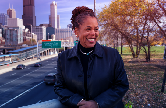 Doctor Beth Richie leaning on a railing smiling to the right in a black jacket and sweater. Chicago skyline and highway to her left and autumn trees to the right.